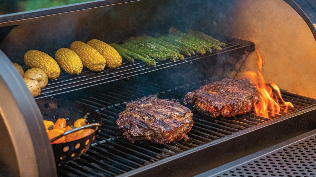 Flames lick steaks searing on the main cooking grate of a grill while vegetables roast on the warming rack