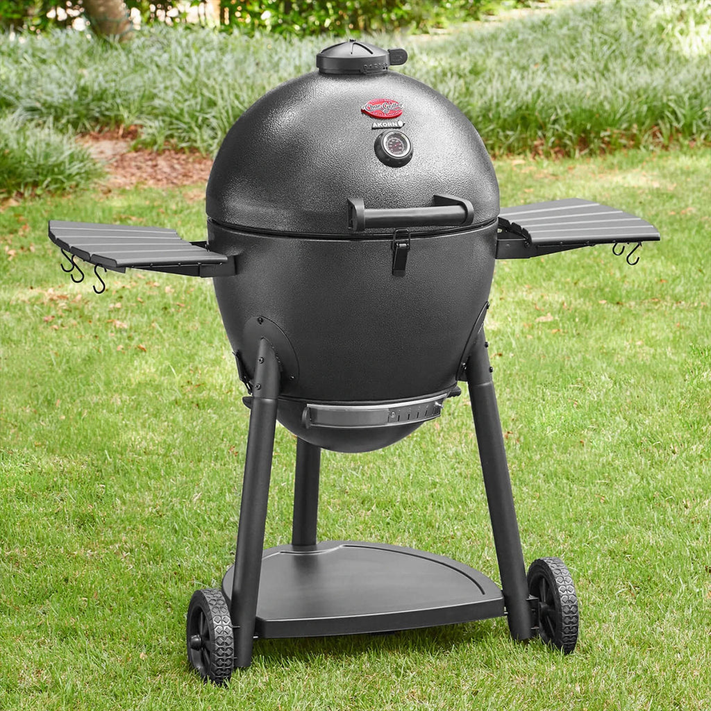 A slate kamado-style grill on a wheeled stand with side shelves, photographed outdoors on a green grass lawn.
