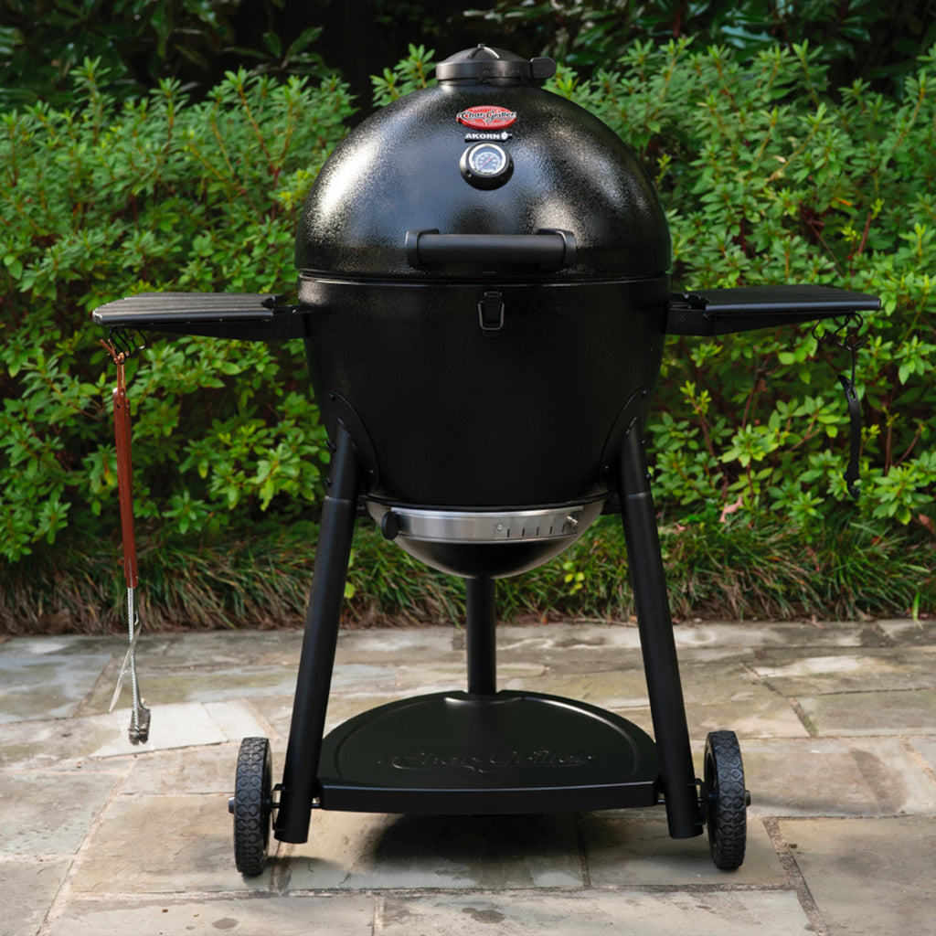 Black AKORN Kamado-style grill on wheels  against a green bush background