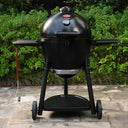 Black AKORN Kamado-style grill on wheels  against a green bush background