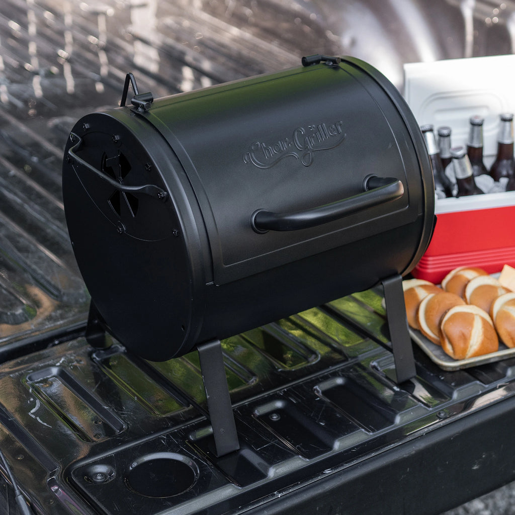 Portable barrel-style grill on the tail-gate of a pickup truck with a tray of  burger buns and a small cooler full of drinks in the background