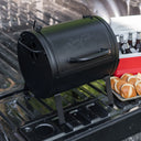 Portable barrel-style grill on the tail-gate of a pickup truck with a tray of  burger buns and a small cooler full of drinks in the background